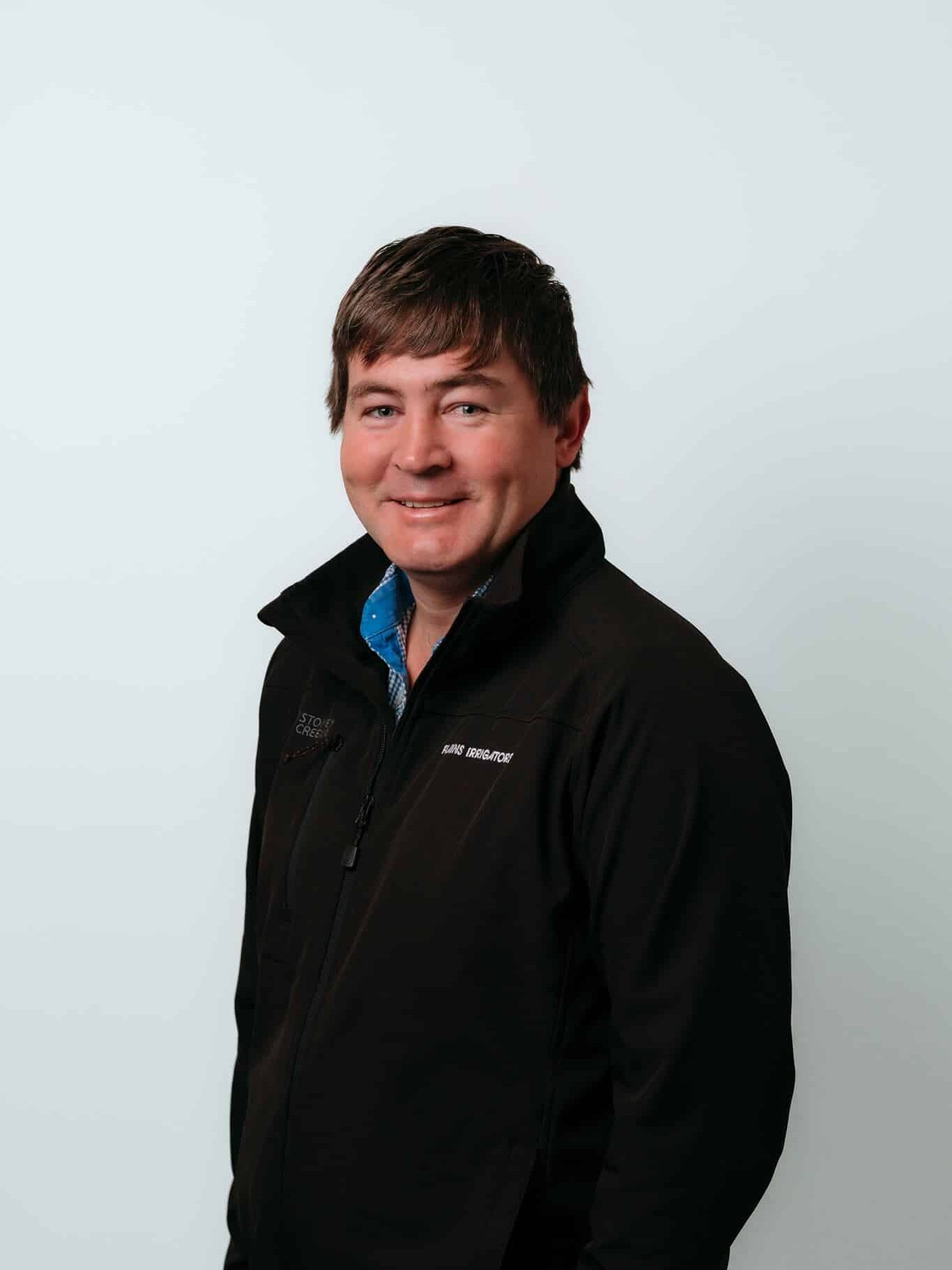 A man with short brown hair, wearing a black jacket and blue collared shirt, stands facing slightly to the right against a plain light background, smiling.