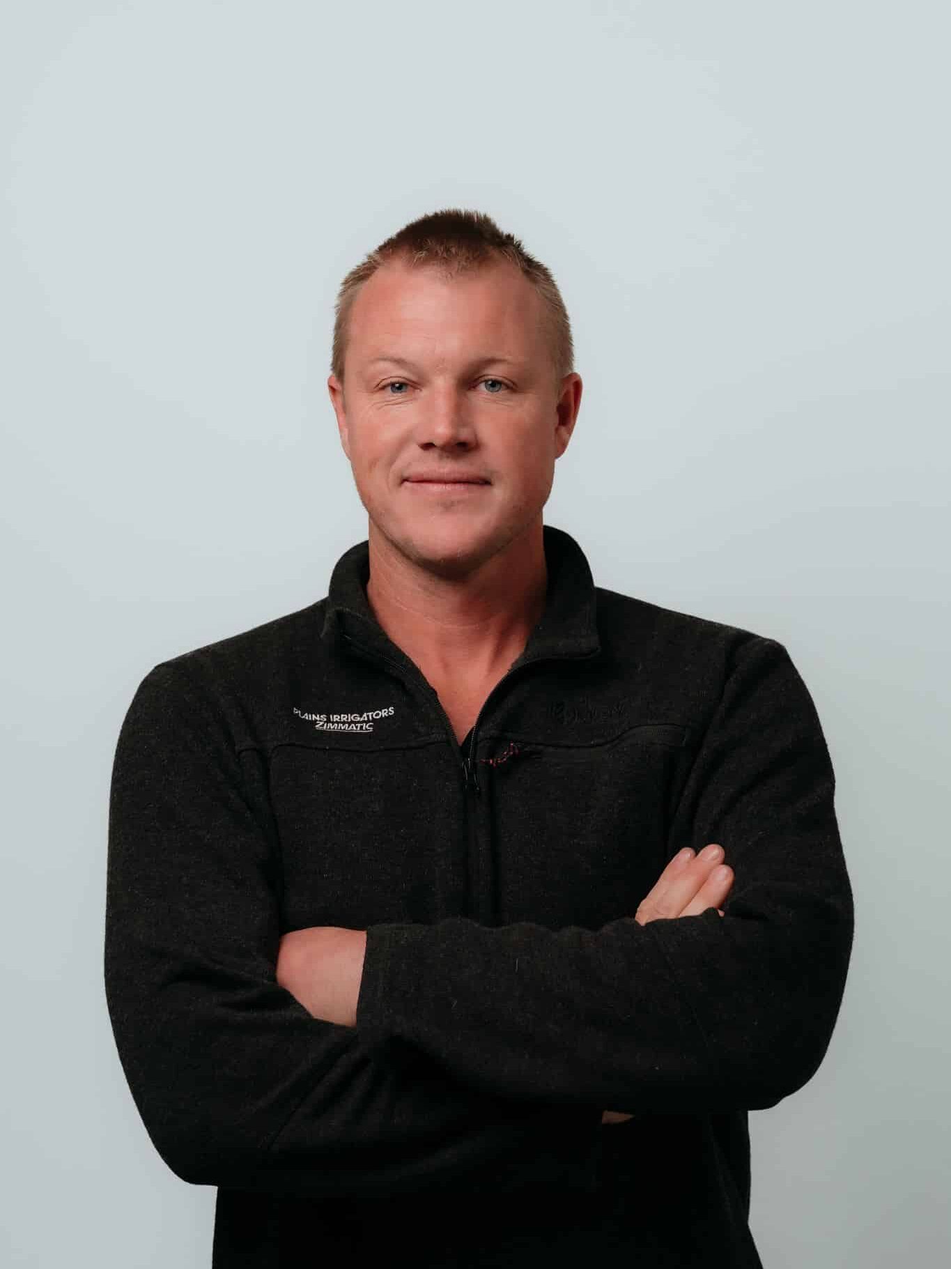 Man with short blond hair wearing a black zip-up sweater stands against a plain light background with arms crossed, looking at the camera with a neutral expression.