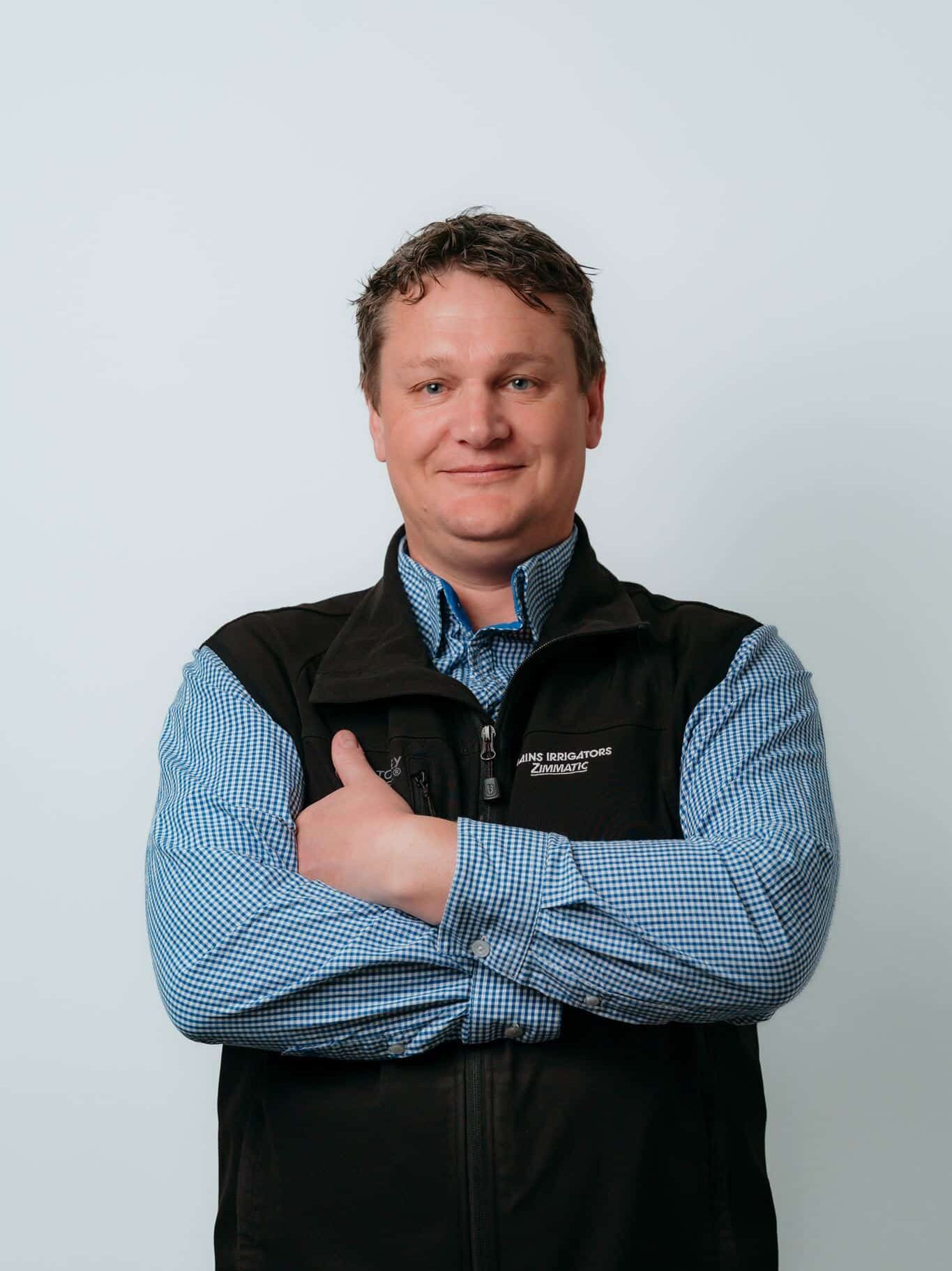 Man in a blue checkered shirt and black vest stands against a plain light background with arms crossed, looking at the camera.