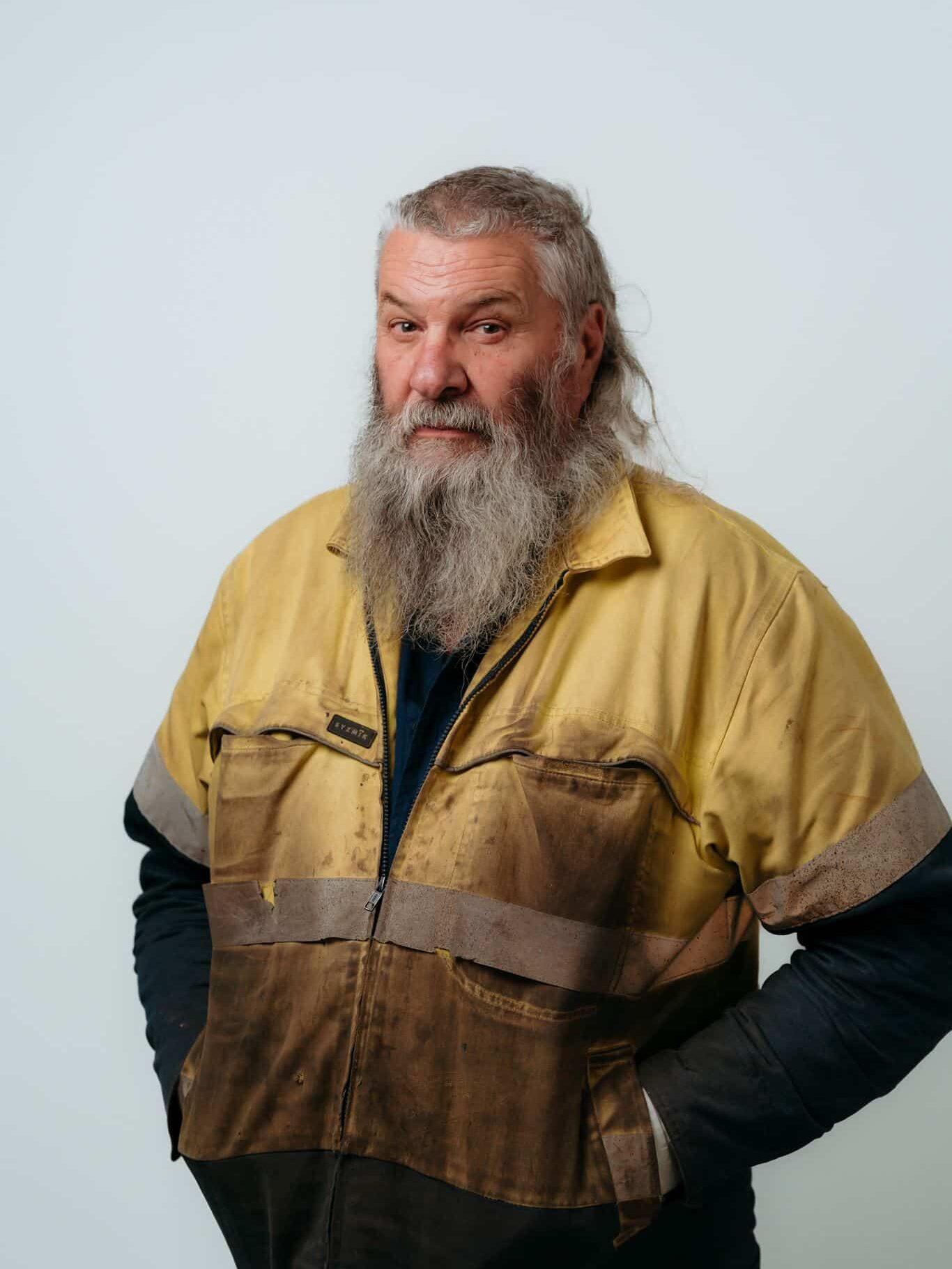 A man with a long gray beard and long hair wears a dirty yellow and brown work jacket, standing against a plain white background with his hands in his pockets.