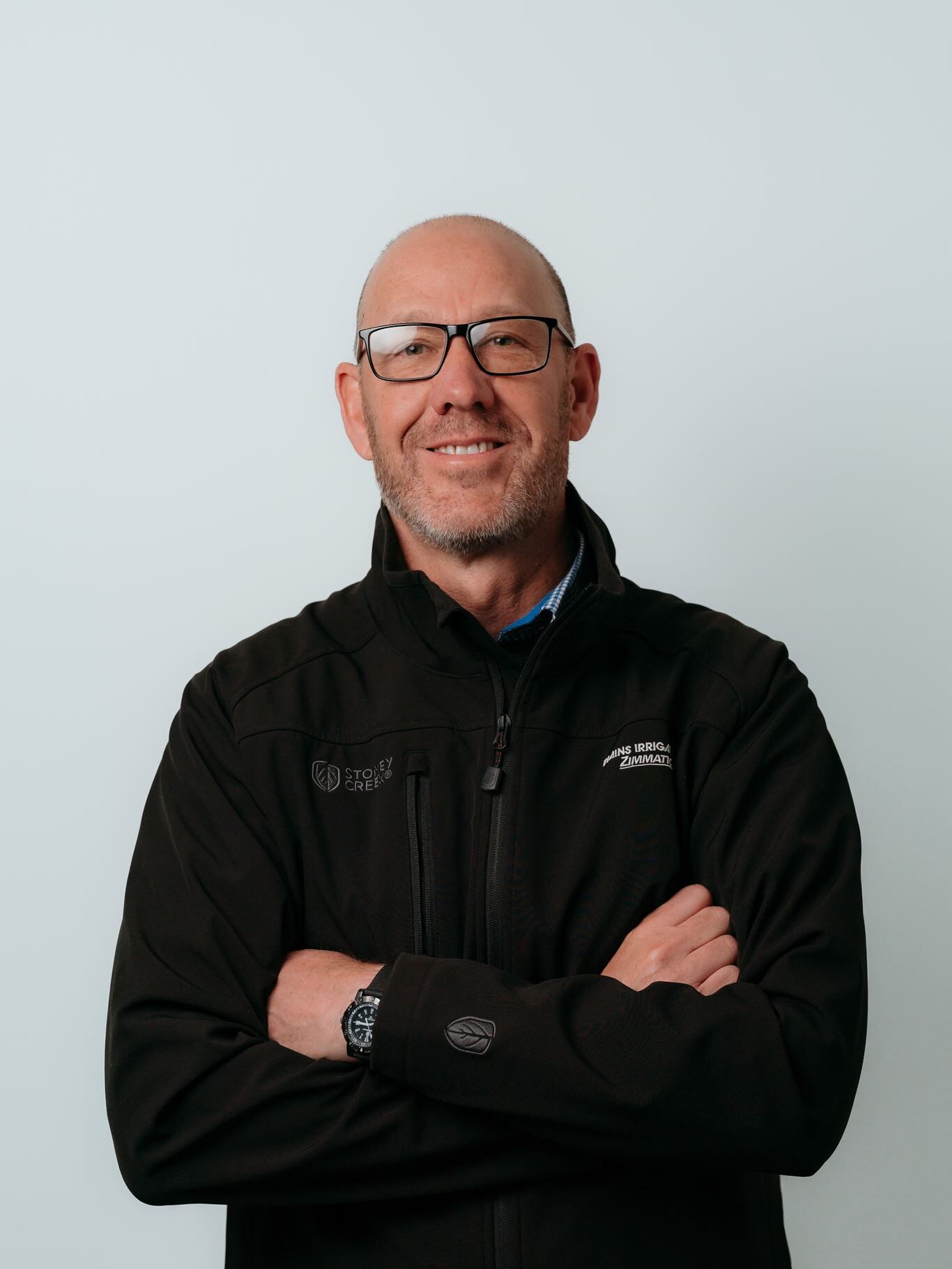 Bald man with glasses wearing a black zip-up jacket stands against a plain light background with arms crossed, smiling slightly at the camera.