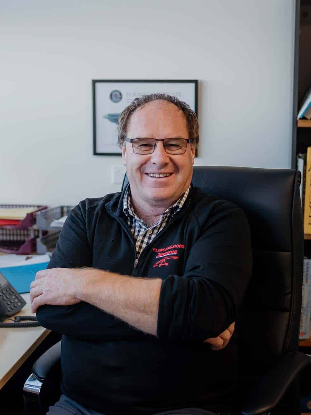 A man sits at a desk in an office, smiling at the camera. His computer monitor displays a spreadsheet and shelves with books and binders are visible in the background.