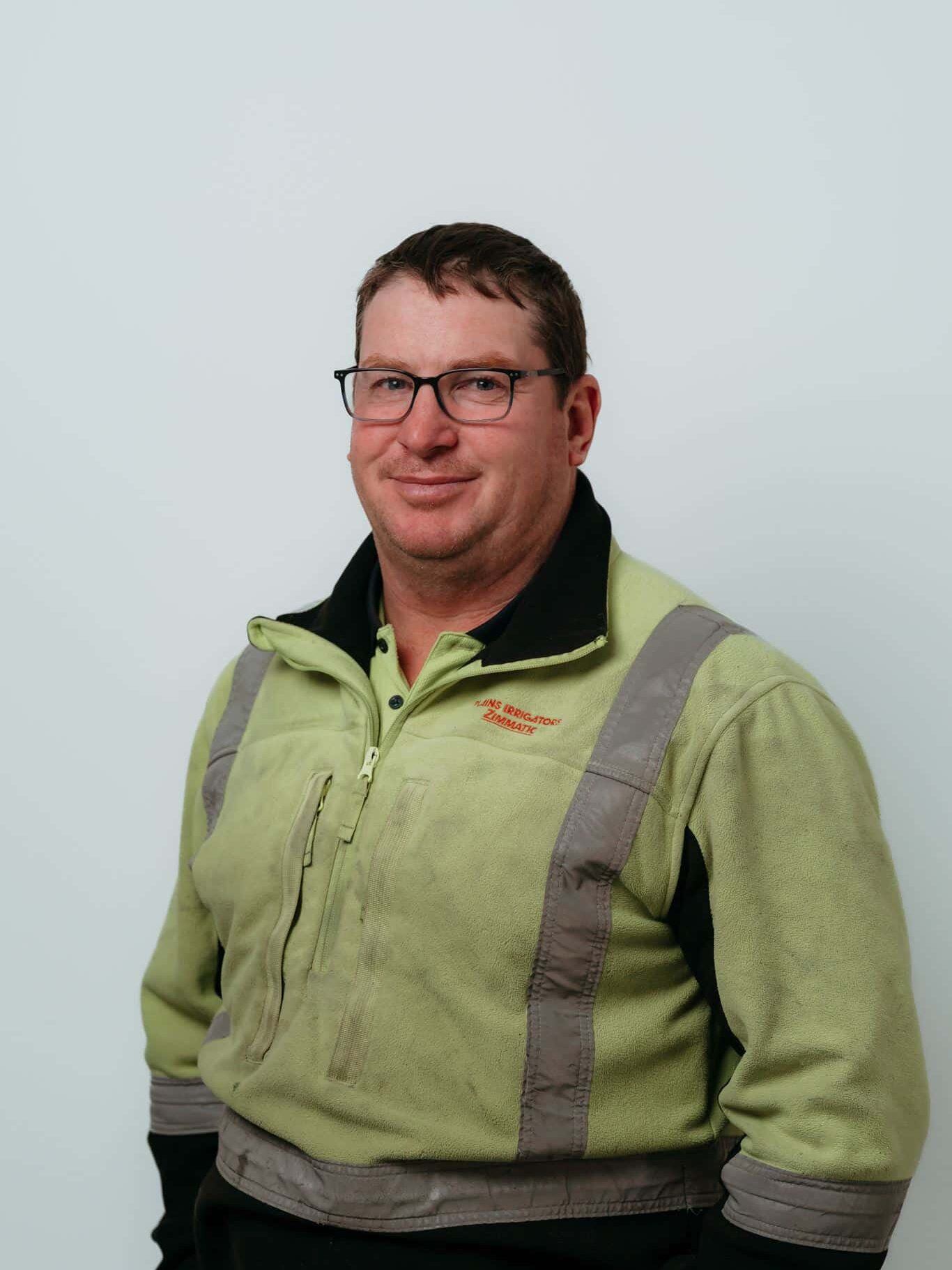 A man in glasses and a green pullover with grey accents stands against a plain light background, facing the camera and smiling slightly.