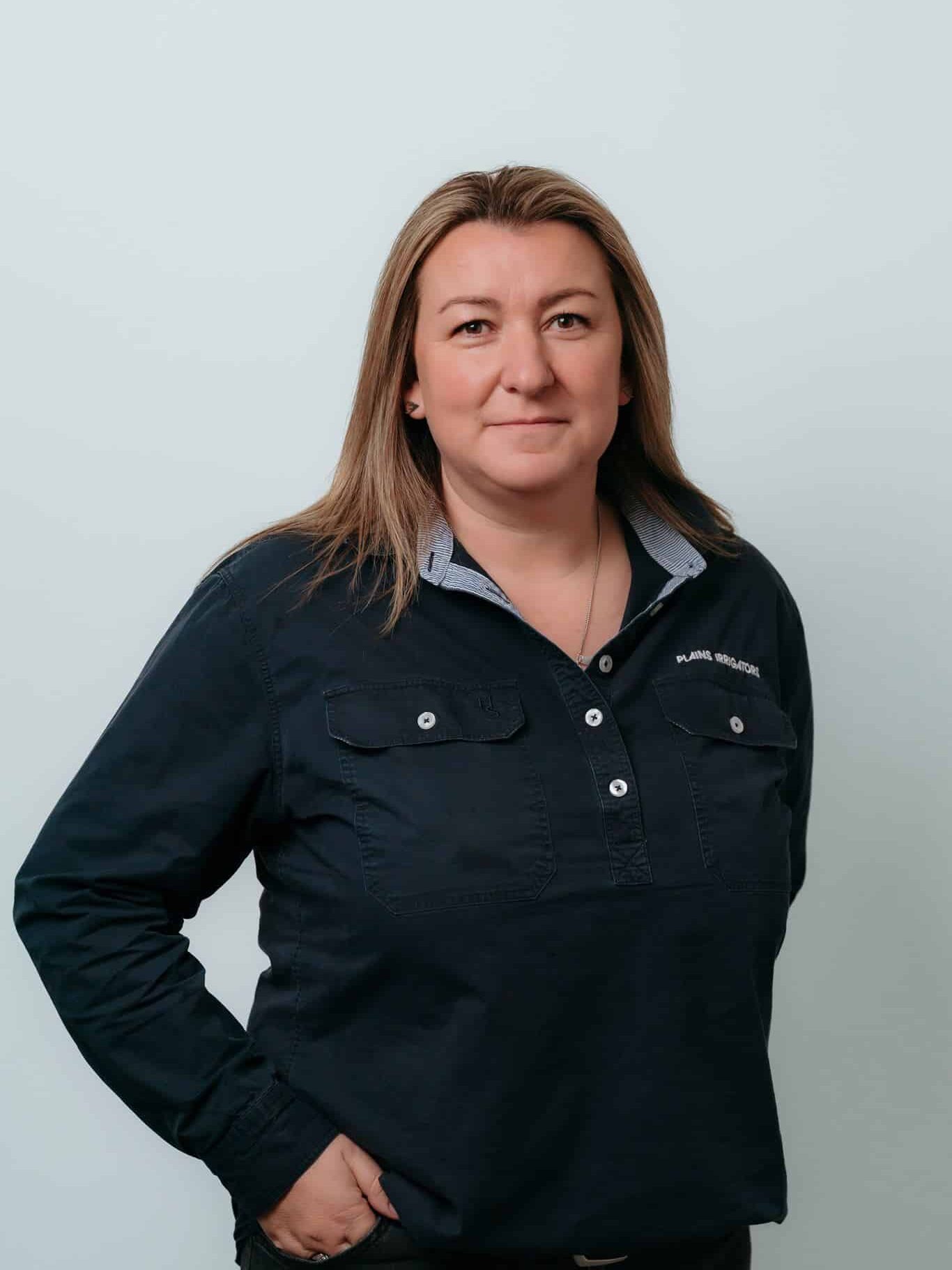 A person with straight, shoulder-length hair wearing a dark button-up shirt stands against a plain light background, looking at the camera with a neutral expression.