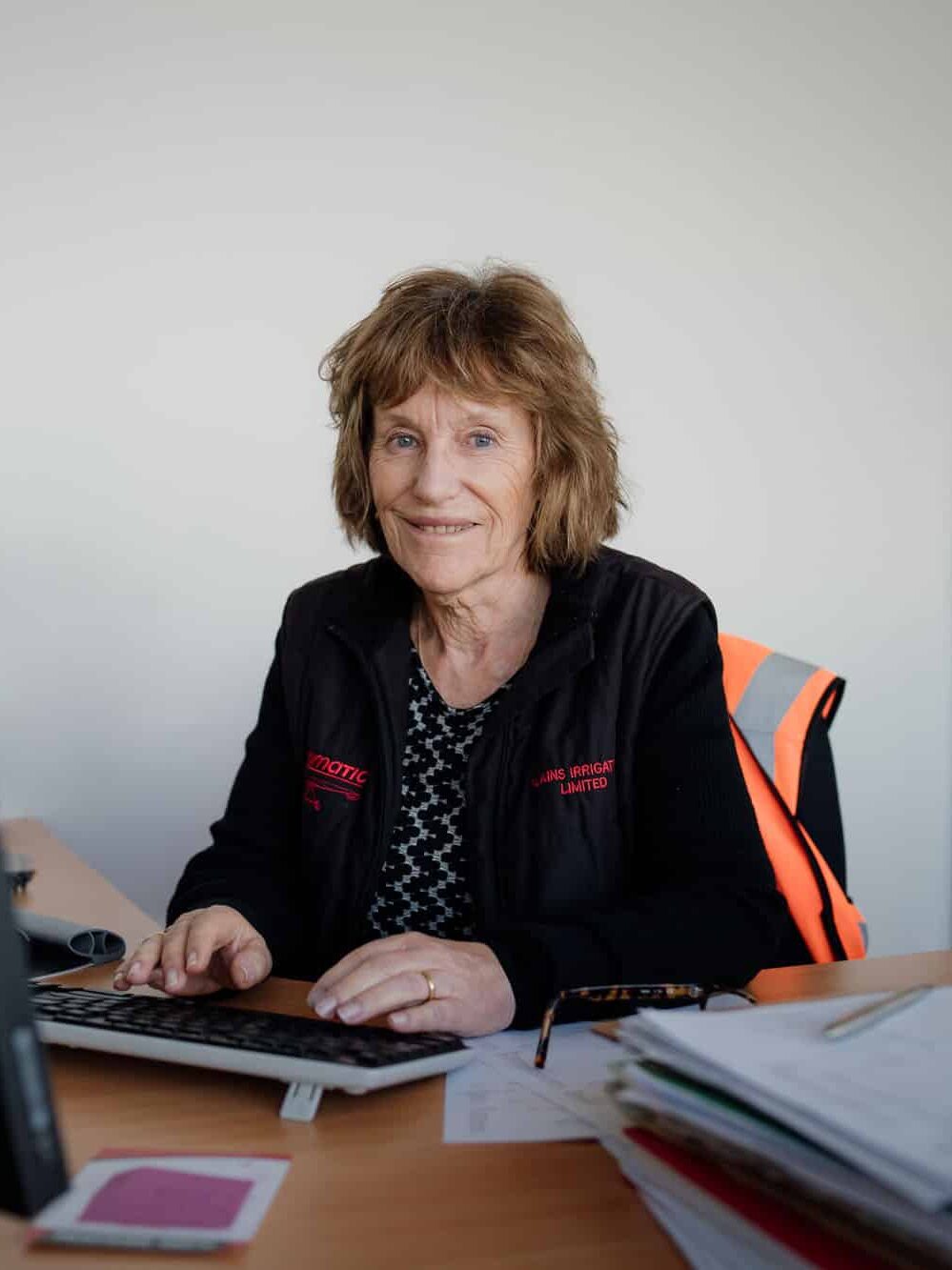 Woman sits at a desk with paperwork, typing on a computer keyboard, in an office with a filing cabinet and smartphone visible.