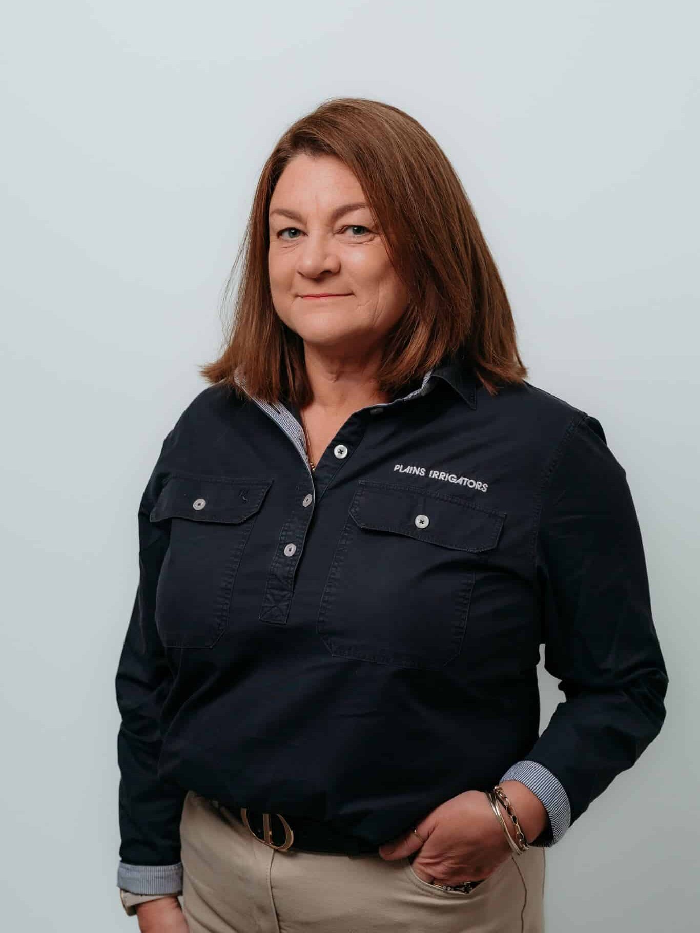 Woman with shoulder-length brown hair stands against a plain light background, wearing a dark collared shirt and khaki pants, with one hand in her pocket.
