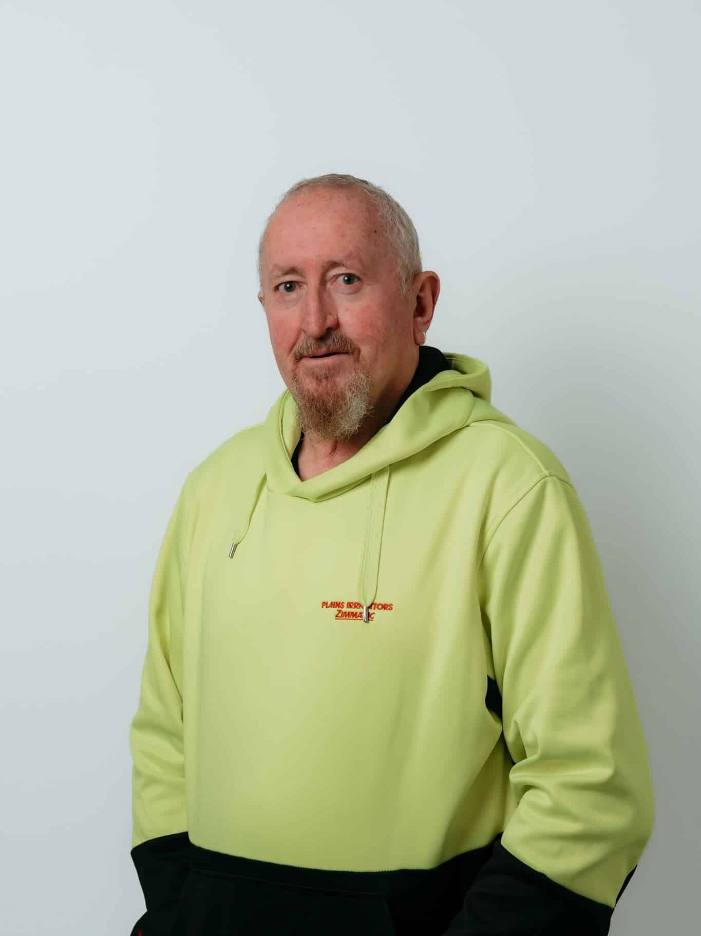 A middle-aged man with short hair and a goatee wears a yellow and black hoodie, standing against a plain white background.